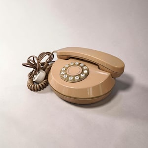 May include: A vintage beige rotary dial telephone with a matching handset. The phone has a circular dial with white numbers and a beige center. The coiled cord and plug are brown.