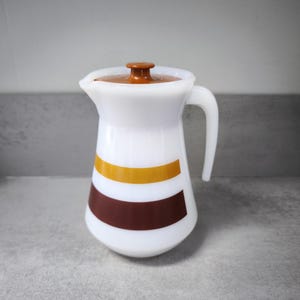 Könnte beinhalten: Eine weiße Kanne mit braunem Deckel und Griff. Die Kanne hat ein braunes und gelbes Streifendesign. Die Kanne ist wahrscheinlich aus Glas oder Keramik und dient zum Servieren von Getränken.
