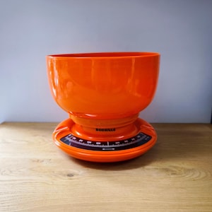 Può includere: Una bilancia da cucina arancione vintage con una grande ciotola e una base circolare. La bilancia presenta un quadrante nero con segni bianchi e il marchio "SOEHNLE". La bilancia è in plastica e poggia su una superficie di legno.