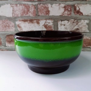 Könnte beinhalten: Eine Keramikschüssel mit glänzendem Finish. Die Schüssel hat eine leuchtend grüne Mitte, die in einen dunkelbraunen Boden und Rand übergeht. Die Schüssel hat eine abgerundete Form und steht auf einem kurzen, stabilen Sockel, der zum Servieren von Speisen geeignet ist.
