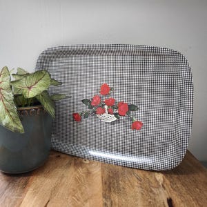 Könnte beinhalten: Rechteckiges Serviertablett mit einem schwarz-weißen Karomuster. Das Tablett hat ein Blumendesign aus roten Rosen und einem weißen Korb. Das Tablett steht auf einer Holzoberfläche, mit einer Topfpflanze im Hintergrund.