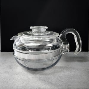 Può includere: Una teiera in vetro trasparente con corpo arrotondato, manico curvo e coperchio. Una fascia color argento circonda la metà della teiera. La teiera è progettata per preparare il tè e ha un beccuccio.