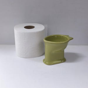 May include: A white toilet paper roll positioned next to a small, olive green ceramic holder shaped like a miniature toilet bowl. The holder features a smooth, glossy finish with a wide opening at the top and a stable, oval-shaped base.