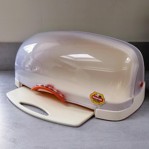 May include: A cream-coloured bread bin with a clear, curved lid and a pull-out cutting board. The bin has an orange handle and a small sticker with text. The design is practical for storing and slicing bread.