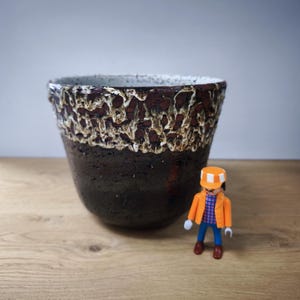 May include: A ceramic planter with a dark brown base and a textured, lighter brown and white rim. A small, orange-jacketed figurine stands beside the planter. The planter has a wide opening and a slightly irregular shape.