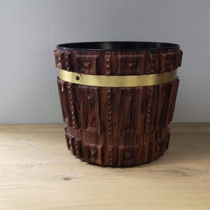 May include: A wooden planter with a brass band around the top. The planter has a dark brown wood grain pattern.