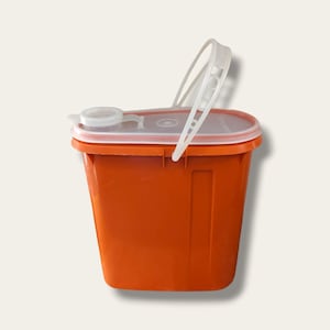 Può includere: Un contenitore arancione per oggetti taglienti con coperchio e manico bianchi. Il contenitore rettangolare ha un coperchio incernierato con una piccola apertura e un manico in plastica trasparente che si incurva sulla parte superiore. Per lo smaltimento sicuro di oggetti taglienti medici.