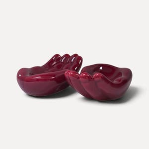 Op de afbeelding: Twee glanzende, diepbordeauxrode keramische schaaltjes in de vorm van komvormige handen. Elk schaaltje heeft een glad, reflecterend oppervlak met gedefinieerde vinger- en handpalmcontouren, ontworpen om kleine voorwerpen in het midden te houden.