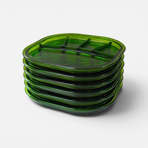 Op de afbeelding: Een stapel van zes vierkante, doorschijnende smaragdgroene glazen borden. Elk bord heeft afgeronde hoeken en een gegoten ontwerp met drie vakken op het oppervlak. Het glas heeft een gladde, glanzende afwerking in een diepgroene kleur.