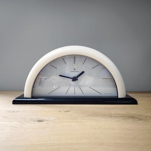 May include: A vintage Junghans clock with a white, arched top and a black rectangular base. The clock face is white with black hands and markers. The clock reads "Junghans" and "Made in Holland".