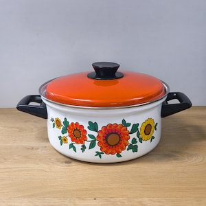Peut inclure: Une marmite en métal émaillé blanc avec un couvercle orange vif et deux poignées latérales noires. L'extérieur de la marmite présente un motif floral décoratif avec de grandes fleurs orange et jaunes et des feuilles vertes autour de la base.