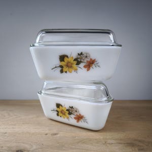 May include: Two white rectangular glass baking dishes with clear glass lids. Each dish features a floral design with yellow, white, and orange flowers. The dishes are stacked on top of each other.