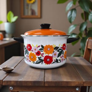 Op de afbeelding: Een witte emaille kookpot met een oranje deksel en zwarte handvatten. De pot is versierd met een bloemenpatroon met rode, oranje en roze bloemen met groene bladeren. De pot staat op een houten tafel.