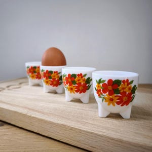Op de afbeelding: Vier witte keramische eierdopjes met een bloemenpatroon in rood, oranje en geel. Eén dopje bevat een bruin ei. De eierdopjes hebben drie kleine voetjes en staan op een houten plank.