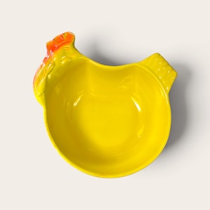Peut inclure: Un bol en céramique jaune vif en forme de poule. Le bol présente une tête sculptée en relief avec une crête et des barbillons orange vif sur le côté gauche, ainsi qu'une section de queue texturée sur le côté droit, formant une silhouette.