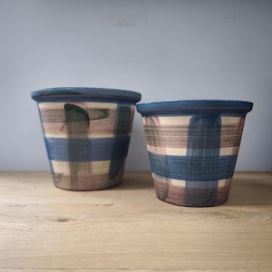 Op de afbeelding: Twee keramische bloempotten met een blauwe rand en een geschilderd ruitpatroon in tinten blauw, beige en groen. De potten zijn cilindrisch en staan op een houten oppervlak. De achtergrond is effen lichtblauw.