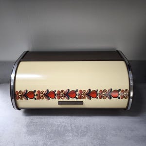 May include: Cream-coloured bread bin with a brown top and a decorative floral pattern in red, brown, and black. The bin has a curved design with chrome accents and a small handle.