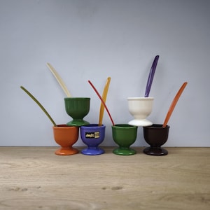 Op de afbeelding: Een verzameling kleurrijke keramische eierdopjes met bijpassende lepels. De eierdopjes zijn in verschillende kleuren, waaronder oranje, groen, blauw, wit en bruin. Elke beker bevat een lepel in een bijpassende kleur. Op de blauwe beker staat de tekst "amfa".