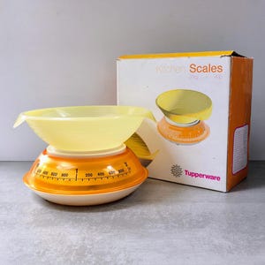 May include: A Tupperware kitchen scale with a translucent yellow bowl and an orange base featuring a measurement dial ranging from 0 to 2 kg and 4 lbs. The scale is displayed next to its original white and orange cardboard packaging box.
