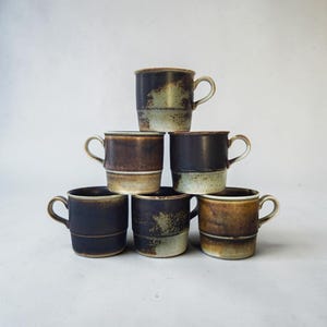 May include: A stack of six ceramic mugs arranged in a pyramid formation. Each mug features a cylindrical body with a side handle and a distinct two-tone glaze, blending dark charcoal, brown, and muted beige tones with a textured, mottled finish.