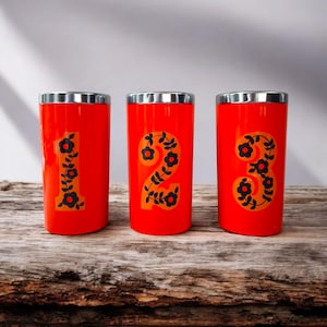 May include: Three red metal cups with silver lids. Each cup has a black and orange floral design with a number, 1, 2, and 3, on the front.