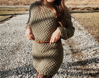 Può includere: Una donna indossa un abito di maglia verde con una cintura in pelle marrone. Indossa un cappello marrone a tesa larga e collant neri con motivo.