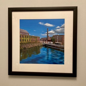 Pu&ograve; includere: Un dipinto incorniciato di un edificio in mattoni con un alto camino, riflesso in un fiume blu. L'edificio &egrave; ricoperto di viti verdi e il cielo &egrave; blu con nuvole bianche.