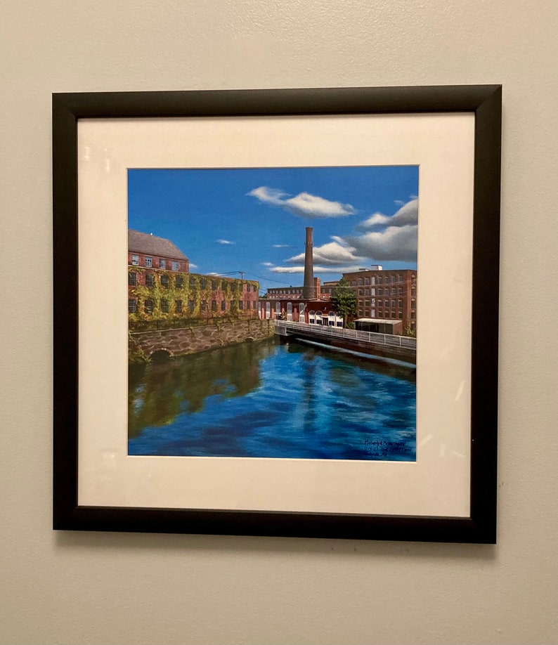 Pu&ograve; includere: Un dipinto incorniciato di un edificio in mattoni con un alto camino, riflesso in un fiume blu. L'edificio &egrave; ricoperto di viti verdi e il cielo &egrave; blu con nuvole bianche.