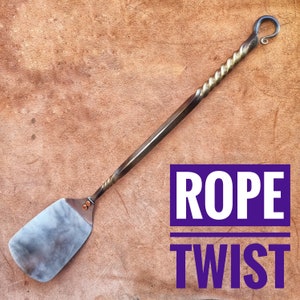 Peut inclure: Spatule en métal forgé à la main, avec une lame plate et rectangulaire et un manche torsadé. La poignée se transforme en un design droit et facetté et se termine par une boucle. Le texte "ROPE TWIST" est dans un encadré violet.