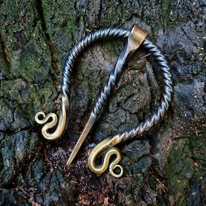May include: A handcrafted metal brooch with a twisted wire design and two decorative loops. The brooch is made of dark metal with a gold-toned finish.