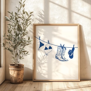 May include: A white framed print with a blue illustration of a clothesline with a bikini, cowboy boots, and a cowboy hat hanging on it.