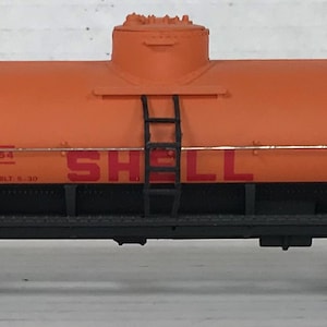 May include: Orange model train tank car with black wheels and a ladder on the side. The car is labeled "SHELL" and "S.C.C.X. 1794".