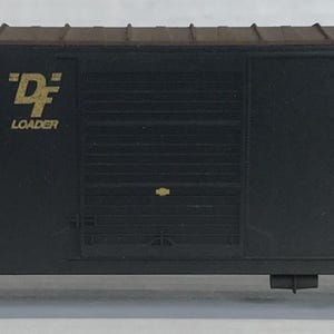 May include: A black model train car with gold lettering and logos. The text on the car reads "ANOTHER CUSHIONED LOAD" and "ATLANTIC COAST LINE". The car has wheels and is designed to look like a real freight car.