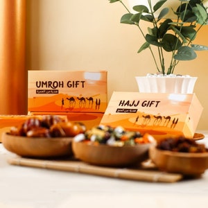May include: Boxes labelled "UMROH GIFT" and "HAJJ GIFT" with desert landscape designs and camel silhouettes. Wooden bowls filled with dates and mixed nuts are in the foreground. A plant in a white pot is in the background.