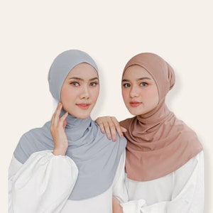 May include: Two women wearing headscarves. One is wearing a light grey head covering, and the other is wearing a tan head covering. Both are wearing white shirts. The background is a light beige colour.