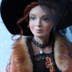 May include: A porcelain doll with red hair and blue eyes, wearing a black hat with a veil. The doll is dressed in a peach-coloured coat with a fur collar, a black lace top, and a beaded necklace with a pendant. The doll has a cross-shaped embellishment on the bodice.