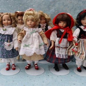 May include: A collection of porcelain dolls, each dressed in detailed, vintage-style outfits. The dolls have painted faces and wear dresses with floral patterns, lace, and ribbons. One doll wears a red hooded cape. The dolls stand on white circular bases.