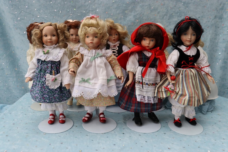 May include: A collection of porcelain dolls, each dressed in detailed, vintage-style outfits. The dolls have painted faces and wear dresses with floral patterns, lace, and ribbons. One doll wears a red hooded cape. The dolls stand on white circular bases.