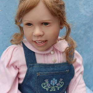May include: A doll with light skin, blonde hair, and blue eyes. The doll wears a pink long-sleeved shirt under a denim overall dress. The dress features a pocket with embroidered flowers. The doll has small earrings and a slight smile.