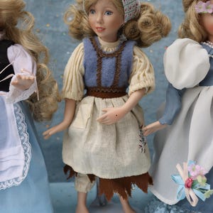 May include: Three porcelain dolls in period costumes. The central doll wears a blue vest over a cream-colored blouse and a tan skirt. The doll on the left wears a blue dress with a white apron. The doll on the right wears a blue dress with a white shawl.