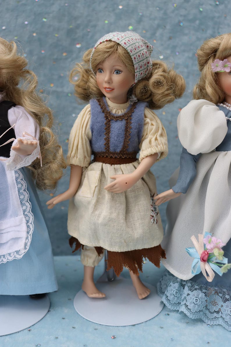 May include: Three porcelain dolls in period costumes. The central doll wears a blue vest over a cream-colored blouse and a tan skirt. The doll on the left wears a blue dress with a white apron. The doll on the right wears a blue dress with a white shawl.