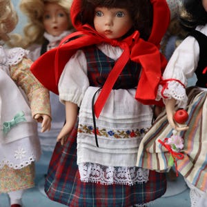 May include: A collection of porcelain dolls dressed in traditional folk costumes. The central doll wears a red hooded cape, a plaid dress, and a white apron with floral embroidery. Other dolls are visible, each in unique outfits.