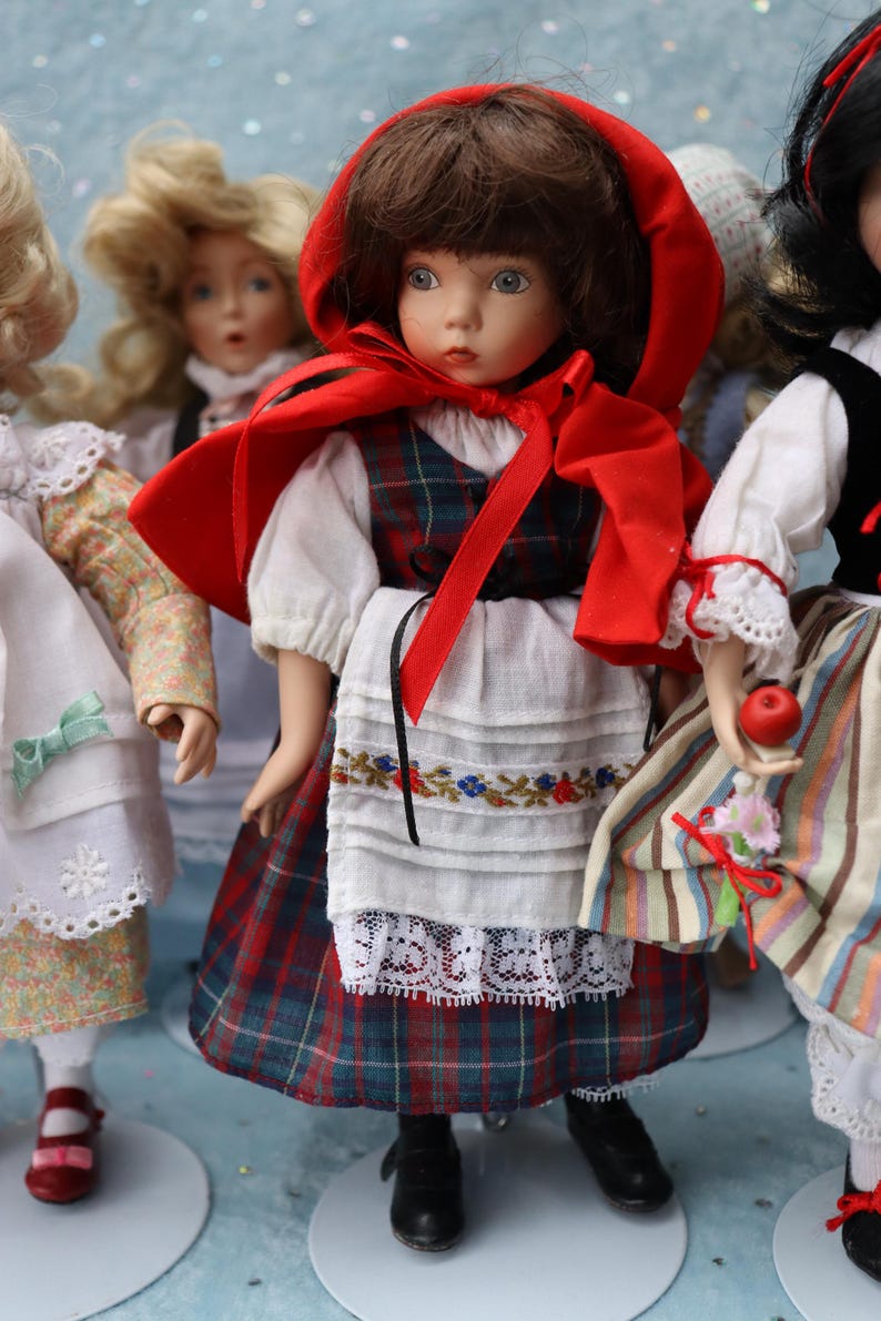 May include: A collection of porcelain dolls dressed in traditional folk costumes. The central doll wears a red hooded cape, a plaid dress, and a white apron with floral embroidery. Other dolls are visible, each in unique outfits.