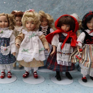 May include: A collection of porcelain dolls, each dressed in detailed, vintage-style outfits. The dolls have painted faces and are posed on small, round stands. The outfits include floral prints, lace, and plaid patterns, with red and black shoes.