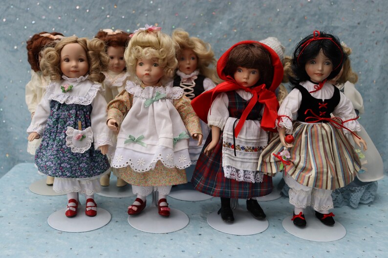 May include: A collection of porcelain dolls, each dressed in detailed, vintage-style outfits. The dolls have painted faces and are posed on small, round stands. The outfits include floral prints, lace, and plaid patterns, with red and black shoes.