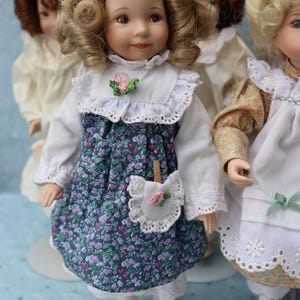 May include: A collection of porcelain dolls. The central doll wears a white blouse, a blue floral dress, and red shoes. The dress has a white lace collar and a small pocket with a pink rose. Other dolls are visible in the background.