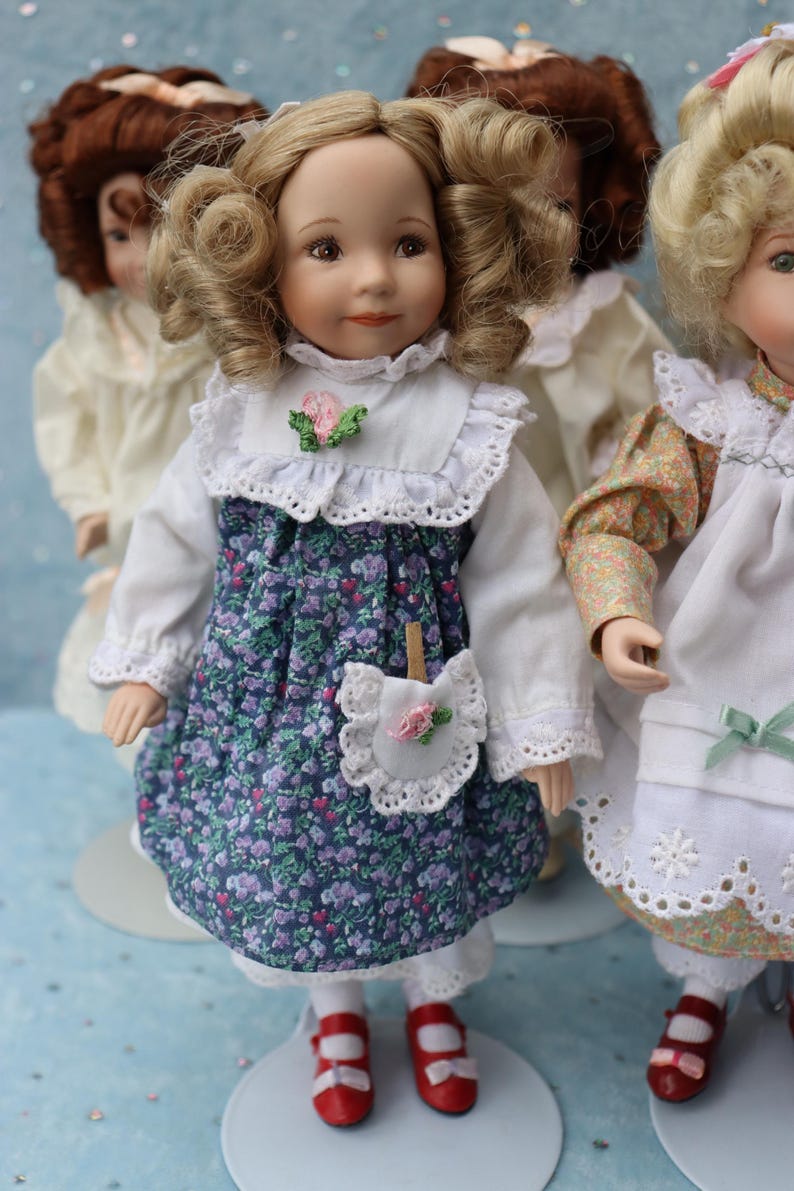 May include: A collection of porcelain dolls. The central doll wears a white blouse, a blue floral dress, and red shoes. The dress has a white lace collar and a small pocket with a pink rose. Other dolls are visible in the background.