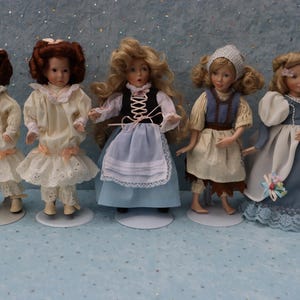 May include: Five porcelain dolls in various dresses and hairstyles stand on white circular bases. The dolls have detailed facial features and wear dresses in shades of white, blue, and brown. The dolls are approximately 8 inches tall.