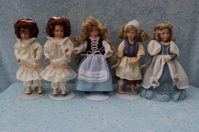 May include: Five porcelain dolls in various dresses and hairstyles stand on white circular bases. The dolls have detailed facial features and wear dresses in shades of white, blue, and brown. The dolls are approximately 8 inches tall.