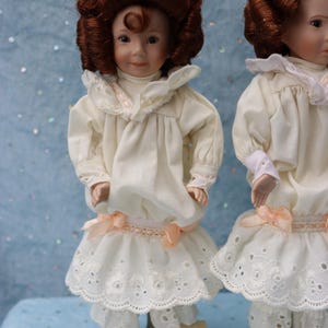 May include: Two porcelain dolls dressed in cream-colored outfits with lace trim and peach-colored bows. The dolls have brown curly hair and are standing on a light blue surface. The dolls are wearing matching outfits, including dresses and pants.